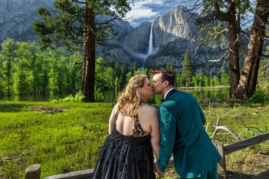 A bride wearing an untraditional black wedding gown smooches her husband in front of Yosemite Falls in this colorful yosemite elopement photo.