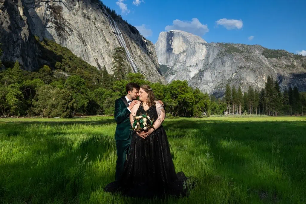 A bride wearing a non traditional black dress cuddles up to her husband in the meadow in front of half dome in their yosemite elopement photos.