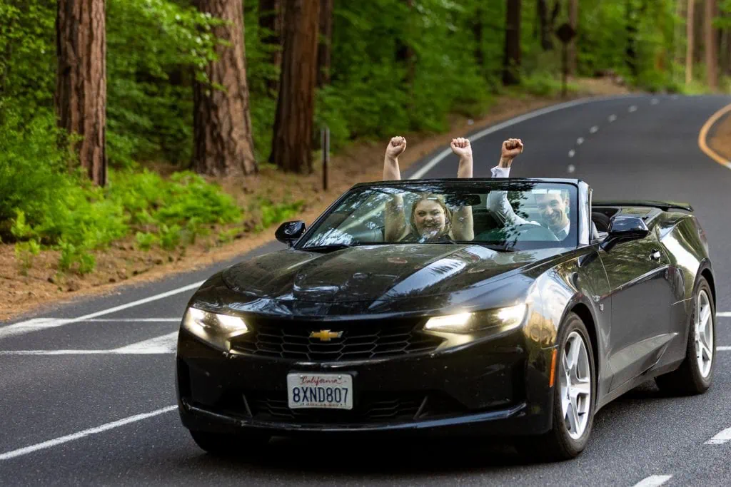 The newlywed bride and groom pump their fists in the sky as they drive away in their black convertible.
