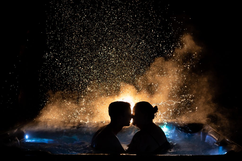 Steam and water droplets rise from behind the silhouetted elopement couple as they bathe in a hot tub at night.