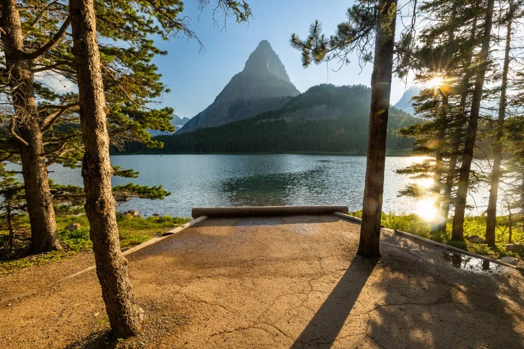 A dirt path leads between trees to a view of Mt Grinnell over Swiftcurrent lake in Glacier National Park.