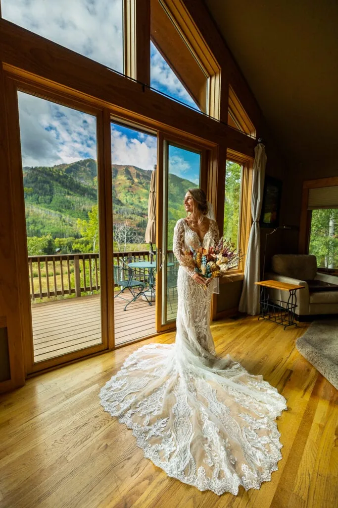 A bride in a long white dress looks out the windows of a beautiful cabin Airbnb in Colorado.
