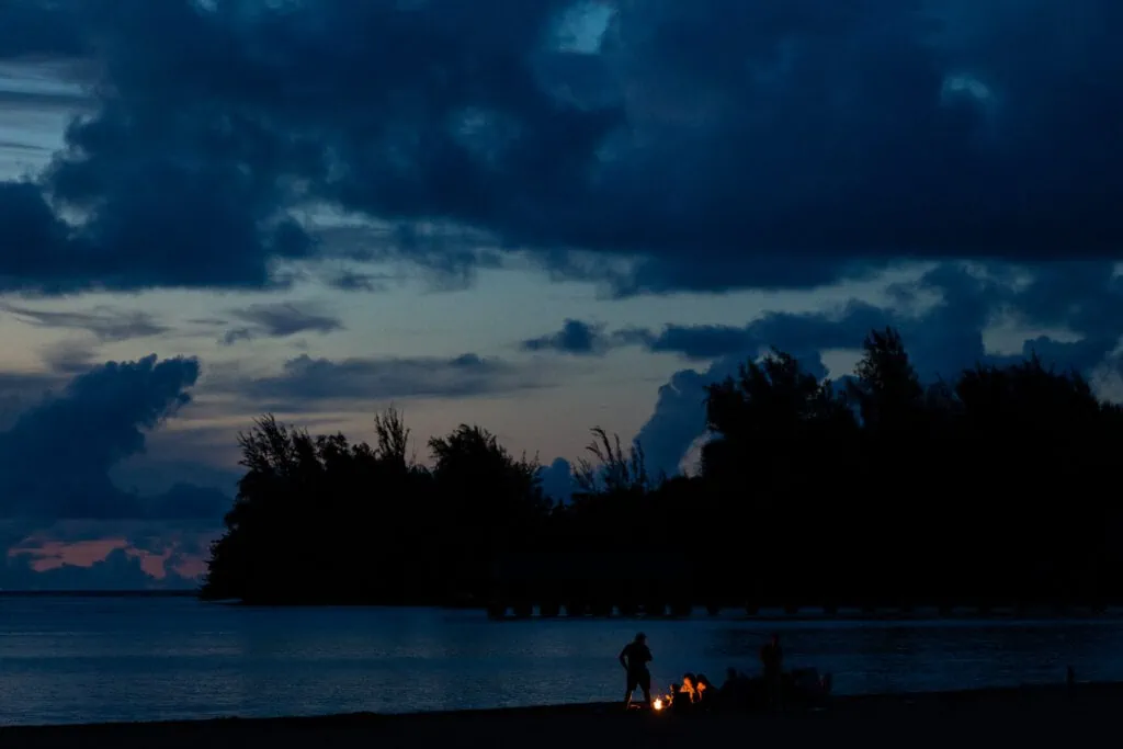 A group of friends gathers around a fire on the beach in Hawaii as the sunset turns into blue twilight.